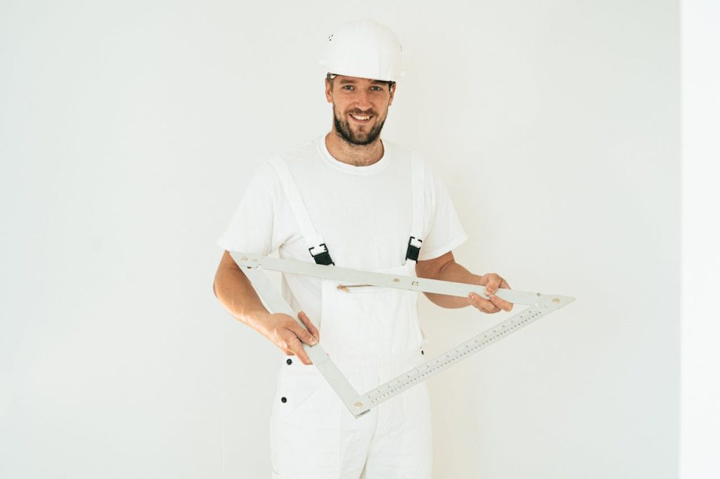 Male builder in white uniform holding a square ruler, wearing a safety helmet against a plain background
