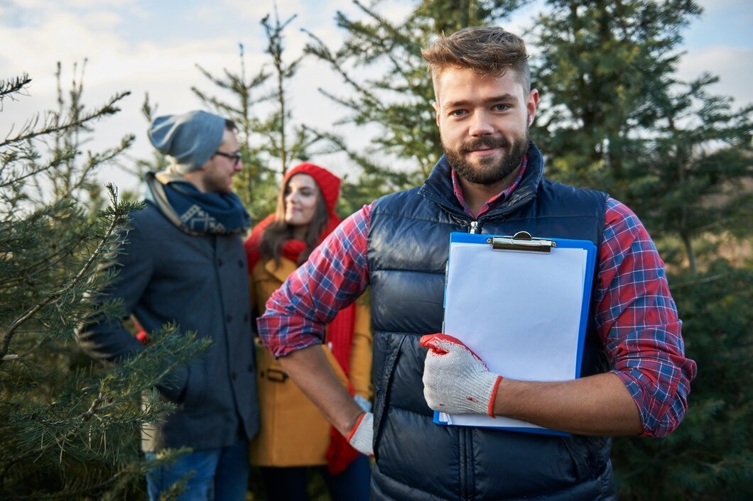 A man stands in front of trees, holding a clipboard and looking thoughtfully at his notes